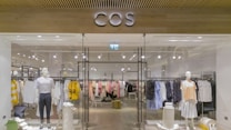 A clothing store display featuring mannequins dressed in casual attire, such as striped and checked shirts and skirts. The store interior is organized with neatly arranged racks of various clothing items, including shirts, blouses, and jackets in a range of colors such as yellow, white, pink, black, and blue. The entrance has glass doors, and the store name is prominently displayed above in bold letters. The lighting is bright, highlighting the clean, modern aesthetic of the space.