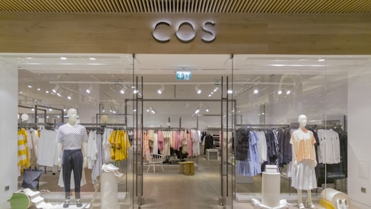 A clothing store display featuring mannequins dressed in casual attire, such as striped and checked shirts and skirts. The store interior is organized with neatly arranged racks of various clothing items, including shirts, blouses, and jackets in a range of colors such as yellow, white, pink, black, and blue. The entrance has glass doors, and the store name is prominently displayed above in bold letters. The lighting is bright, highlighting the clean, modern aesthetic of the space.