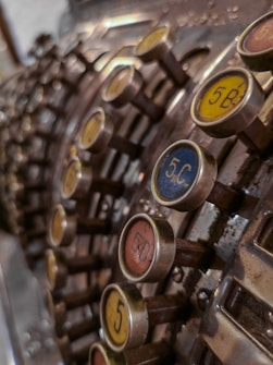A close-up view of an old, mechanical cash register with numerous round buttons displaying numbers and letters in various colors. The buttons are arranged in rows and columns, mounted on long metal rods which protrude at different angles. The surface of the cash register shows signs of wear and age with a metallic, slightly rusty texture.
