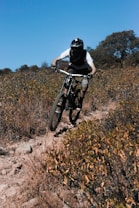 A person wearing protective gear and a helmet is riding a mountain bike on a rough and rocky trail surrounded by dry bushes and shrubs. The rider appears to be taking an aggressive posture, leaning forward while gripping the handlebars tightly. The background features scattered trees under a clear blue sky, showcasing a rugged outdoor environment.