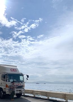 A large white truck with a red logo parked next to a waterfront. A person stands near the truck, holding a beverage. Several small ships or ferries are visible in the distance on a calm ocean. The sky is partly cloudy, with patches of blue and scattered white clouds.