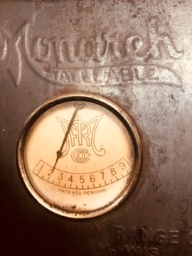 An antique dial or gauge is mounted on a surface, displaying numbers from one to nine with the label 'Patents Pending'. The visible part says 'Monarch' and 'All State'. The surface is metallic and aged, with a rustic appearance.