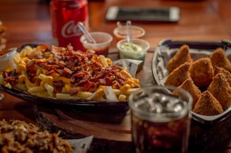 A platter of loaded fries topped with bacon and cheese is accompanied by a tray of golden-brown fried snacks. Various dipping sauces, a can of soda, and a glass with ice are also visible. The scene suggests a hearty and indulgent meal.