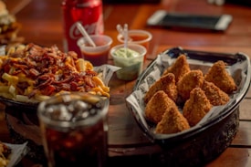 A table setting featuring a tray of crispy, golden-brown, triangular fried snacks and a plate loaded with cheesy fries topped with bacon bits. In the background, there is a red soda can and small cups of sauces.