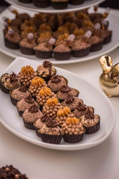 A white oval platter is filled with an assortment of small, intricately decorated chocolate confections. They are arranged in neat rows, featuring varying designs with some having a dome of small round balls and others topped with delicate swirls and floral patterns. The background includes more chocolates on a tiered tray and a gold decorative object.