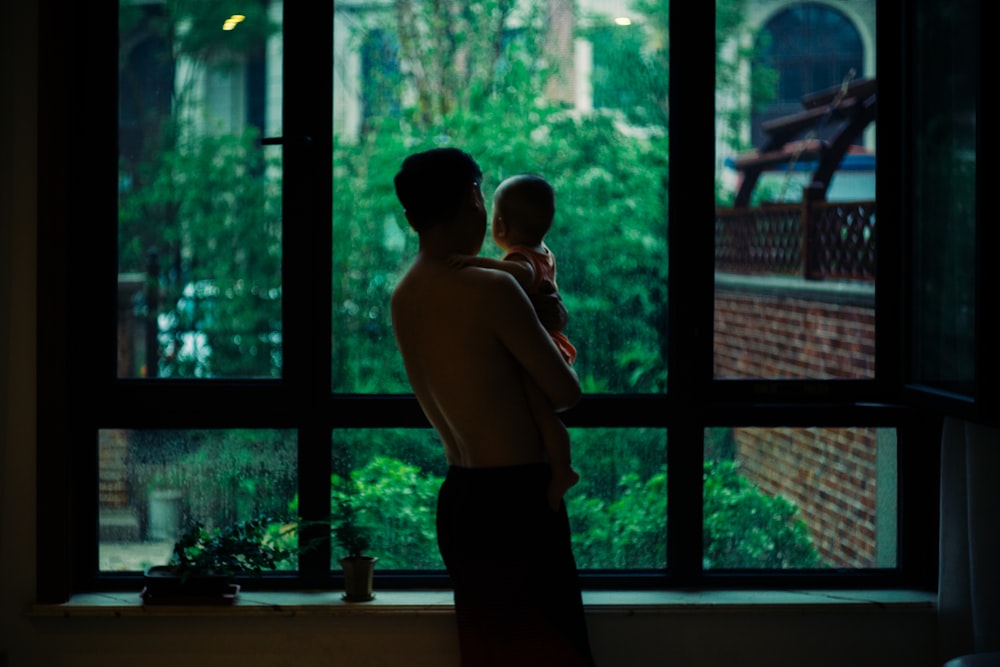 hombre en topless que lleva a un bebé de pie junto a la ventana