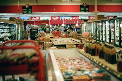 The interior of a supermarket with aisles lined with products such as beverages, frozen goods, and packaged food. Several aisles of refrigerated sections and shelves filled with products are visible. There are signs above each aisle indicating the sections, and a few customers are browsing and shopping.