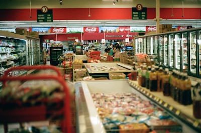 The interior of a supermarket with aisles lined with products such as beverages, frozen goods, and packaged food. Several aisles of refrigerated sections and shelves filled with products are visible. There are signs above each aisle indicating the sections, and a few customers are browsing and shopping.