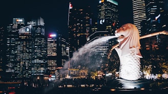 A night scene captures a prominent Merlion statue by the waterfront, spouting water with the backdrop of a brightly lit cityscape. Tall skyscrapers adorned with various illuminated logos form the horizon, creating a vibrant urban atmosphere.