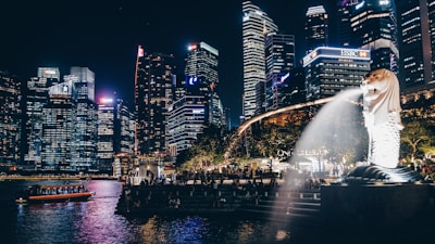 A cityscape at night featuring illuminated skyscrapers next to a body of water. In the foreground, an iconic Merlion statue with water spouting from its mouth is visible, surrounded by trees and people walking by.