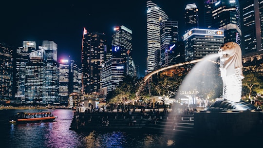 A cityscape at night featuring illuminated skyscrapers next to a body of water. In the foreground, an iconic Merlion statue with water spouting from its mouth is visible, surrounded by trees and people walking by.