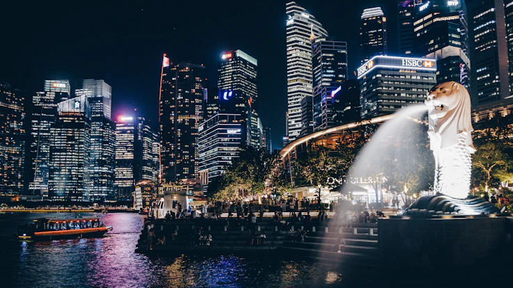 A bright, lively photo of the Singapore skyline at dusk featuring iconic landmarks and a mix of multi-racial Singaporean professionals walking along the riverside.