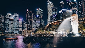 A cityscape at night featuring illuminated skyscrapers next to a body of water. In the foreground, an iconic Merlion statue with water spouting from its mouth is visible, surrounded by trees and people walking by.