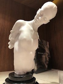 A white marble sculpture depicting a human figure with a tilted head and delicate features, set against a wooden background. The sculpture is mounted on a black, circular base. Behind the sculpture, there is a book with the words 'Richard MacDonald Sculptor' visible.