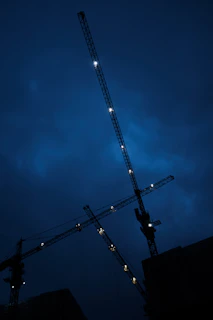 A modern construction site at dusk with cranes and building frames silhouetted against a dark blue sky.