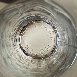 A view looking down into a transparent glass containing embossed text 'OPEN YOUR WORLD' on the bottom. The glass surface shows reflections and concentric circular patterns.