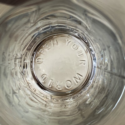 A view looking down into a transparent glass containing embossed text 'OPEN YOUR WORLD' on the bottom. The glass surface shows reflections and concentric circular patterns.