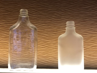 Two glass bottles stand side by side against a textured background. The bottle on the left is clear glass, revealing its transparent quality, while the bottle on the right is frosted, giving it an opaque, milky appearance. The light shines on both bottles from below, highlighting their shapes and details.