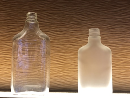 Two glass bottles stand side by side against a textured background. The bottle on the left is clear glass, revealing its transparent quality, while the bottle on the right is frosted, giving it an opaque, milky appearance. The light shines on both bottles from below, highlighting their shapes and details.