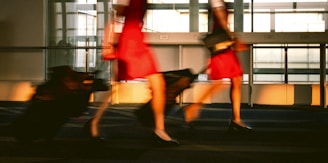 The image captures a motion blur of flight attendants in red uniforms walking briskly through an airport terminal, pulling black rolling luggage. The setting is modern with large glass windows allowing natural light to flood in, casting shadows on the floor.