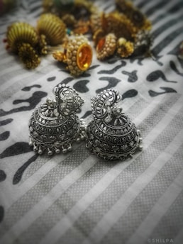 Intricately designed silver jhumka earrings are placed on a patterned surface. The earrings have elaborate detailing and small bell-like structures at the bottom. In the background, several pieces of jewelry with gold, pearls, and stones are displayed, slightly out of focus.