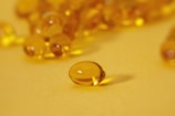 A close-up of several translucent, golden-yellow capsules scattered across a smooth, bright yellow surface. Each capsule is oval-shaped with a shiny, reflective exterior that catches the light. One capsule is prominently in focus at the front, while others appear blurred in the background.