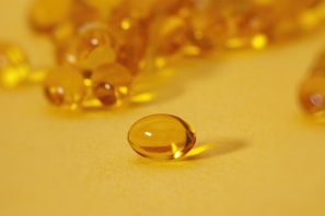 A close-up of several translucent, golden-yellow capsules scattered across a smooth, bright yellow surface. Each capsule is oval-shaped with a shiny, reflective exterior that catches the light. One capsule is prominently in focus at the front, while others appear blurred in the background.