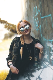 A person with long blonde hair styled in a braid is wearing reflective sunglasses and a camouflage jacket. They are leaning against a wall with graffiti and have an edgy style, including a necklace with a skull pendant.