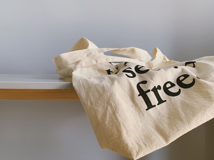 A white cloth tote bag with black text partially visible, resting on the edge of a wooden and white table. The bag's handles are casually draped over its side.
