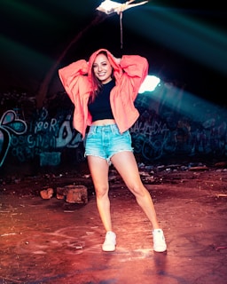 A woman stands confidently in an urban setting with graffiti-covered walls and a grungy floor. She is wearing a vibrant red-orange jacket, denim shorts, and white sneakers. Sunlight streams through an opening above, casting a dramatic light around her.