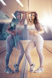 A woman with long hair, wearing a white sports bra and leggings, stands confidently leaning against a glass wall in a gym. Her reflection is visible in the glass, creating a mirror image effect. The gym has wooden floors and bright lighting.
