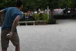 A person in a blue patterned shirt and khaki shorts is playing a game with several silver balls spread across a sandy surface. The person appears to be preparing to throw another ball. The setting is an outdoor park with greenery, benches, and people in the background.