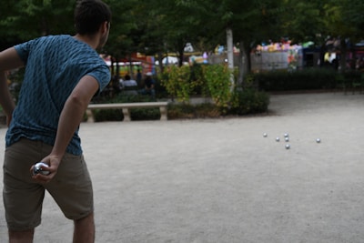 A person in a blue patterned shirt and khaki shorts is playing a game with several silver balls spread across a sandy surface. The person appears to be preparing to throw another ball. The setting is an outdoor park with greenery, benches, and people in the background.