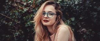 woman in blue-framed glasses standing beside green bushes