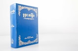 A blue hardcover book with gold ornate designs and text on the cover, standing upright against a plain white background. The text appears to be in an Asian script, likely Khmer.