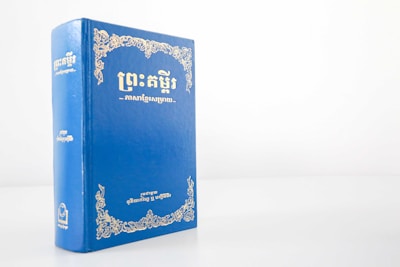 A blue hardcover book with gold ornate designs and text on the cover, standing upright against a plain white background. The text appears to be in an Asian script, likely Khmer.