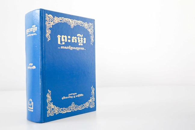 A blue hardcover book with gold ornate designs and text on the cover, standing upright against a plain white background. The text appears to be in an Asian script, likely Khmer.