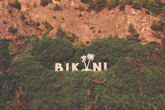 Large white letters spelling 'BIKINI' stand against a backdrop of dense green foliage and trees. The letters are flanked by two palm tree silhouettes. Above, there is a rocky hillside with patches of green bushes and trees.