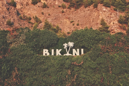 Large white letters spelling 'BIKINI' stand against a backdrop of dense green foliage and trees. The letters are flanked by two palm tree silhouettes. Above, there is a rocky hillside with patches of green bushes and trees.
