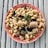 A plate of pasta featuring curly noodles mixed with pieces of sausage, eggplant, and green vegetables, garnished with a basil leaf. The dish is served on a pink plate with a pattern, placed on a rustic wooden surface.