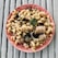 A plate of pasta featuring curly noodles mixed with pieces of sausage, eggplant, and green vegetables, garnished with a basil leaf. The dish is served on a pink plate with a pattern, placed on a rustic wooden surface.