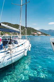 A sailboat is docked in crystal-clear blue water, with a coastal village and forested hills in the background. Other boats are visible further along the shore.