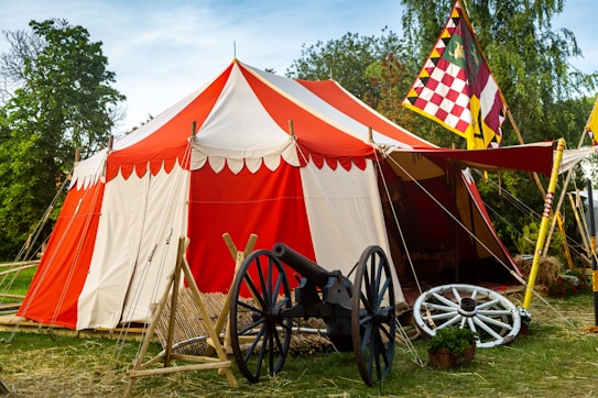 A colorful medieval-style tent with red and white stripes stands prominently in a grassy area. In front of the tent is an old-fashioned cannon and some wooden structures. A decorative flag with a checkerboard pattern in red, yellow, and white is displayed beside the tent. The background shows lush green trees under a clear blue sky.