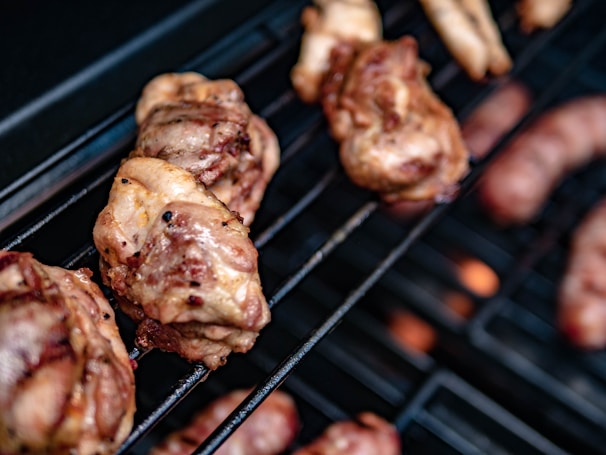Juicy pieces of meat are grilling on a barbecue with visible flames in the background. The meat appears to be well-cooked with grill marks and a slightly crispy outer layer.