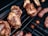Juicy pieces of meat are grilling on a barbecue with visible flames in the background. The meat appears to be well-cooked with grill marks and a slightly crispy outer layer.