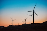 Several wind turbines are positioned on a hill against a vibrant backdrop of a sunset. The sky transitions from a deep blue at the top to a warm orange near the horizon.