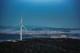 A large wind turbine stands prominently on a hill covered with dense greenery. In the background, an industrial area with cranes and buildings is visible, along with the sea and distant mountains under the evening sky.