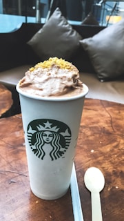 A Starbucks cup filled with a creamy beverage, topped with a generous layer of whipped cream and yellow crumbles, is set on a wooden table. A white plastic spoon and a sugar packet are placed nearby. The background features plush cushions on a sofa.