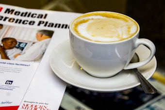 A cup of coffee with frothy milk sits on a saucer accompanied by a teaspoon. Next to it is a magazine or brochure titled 'Medical Conference Planning' featuring an image of two people in conversation, likely in a professional or clinical setting.