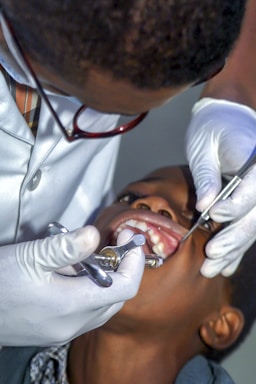 A dentist wearing glasses and gloves is performing a dental procedure on a child. The dentist is using dental instruments, and the child's mouth is open.
